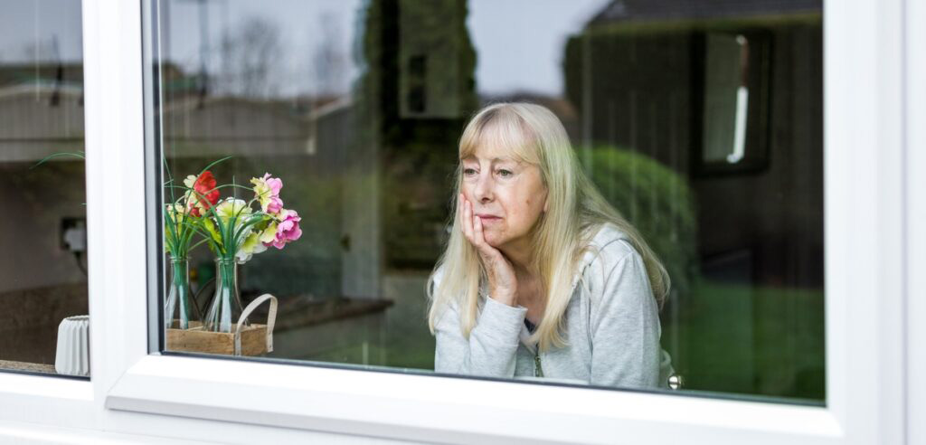 elder abuse attorney, Close up desaturated color image depicting a sad, depressed-looking senior woman in her 70s, and of caucasian ethnicity, looking of her kitchen window at home